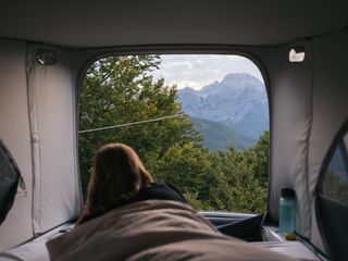 Eine Frau blickt beim Wildcampen in Österreich aus der geöffneten Heckklappe eines Fensters auf eine bewaldete Berglandschaft.